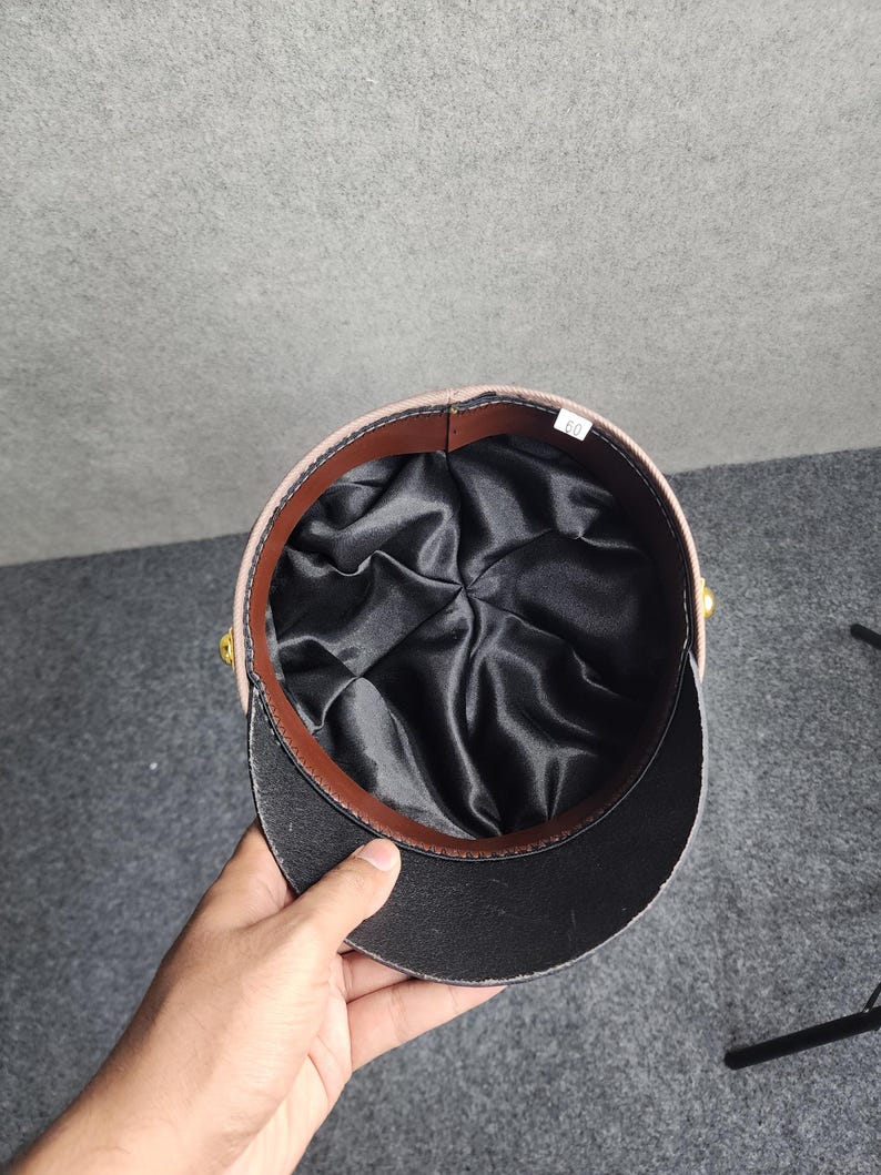 May include: A tan and black military-style hat with a black brim and a brown leather band. The interior is lined with black satin. The hat has gold-colored accents on the sides. The hat is being held by a hand.