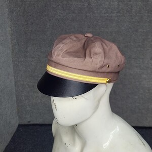 May include: A brown newsboy cap with a black brim and a gold-colored band. The cap is displayed on a white mannequin head against a gray background. The cap has a button on top and small gold-colored eyelets.