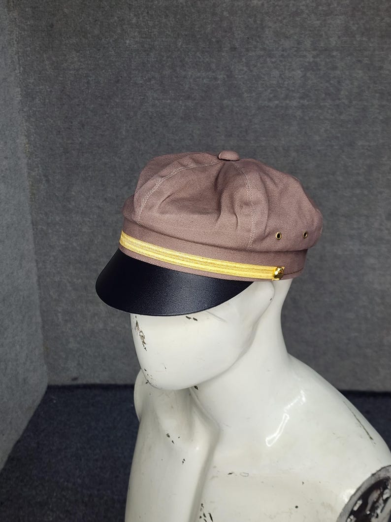 May include: A brown newsboy cap with a black brim and a gold-colored band. The cap is displayed on a white mannequin head against a gray background. The cap has a button on top and small gold-colored eyelets.