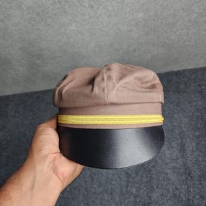 May include: A brown cap with a black brim and a yellow band. The cap is held in a hand against a gray background. The cap has a rounded top and a stiff, curved brim.
