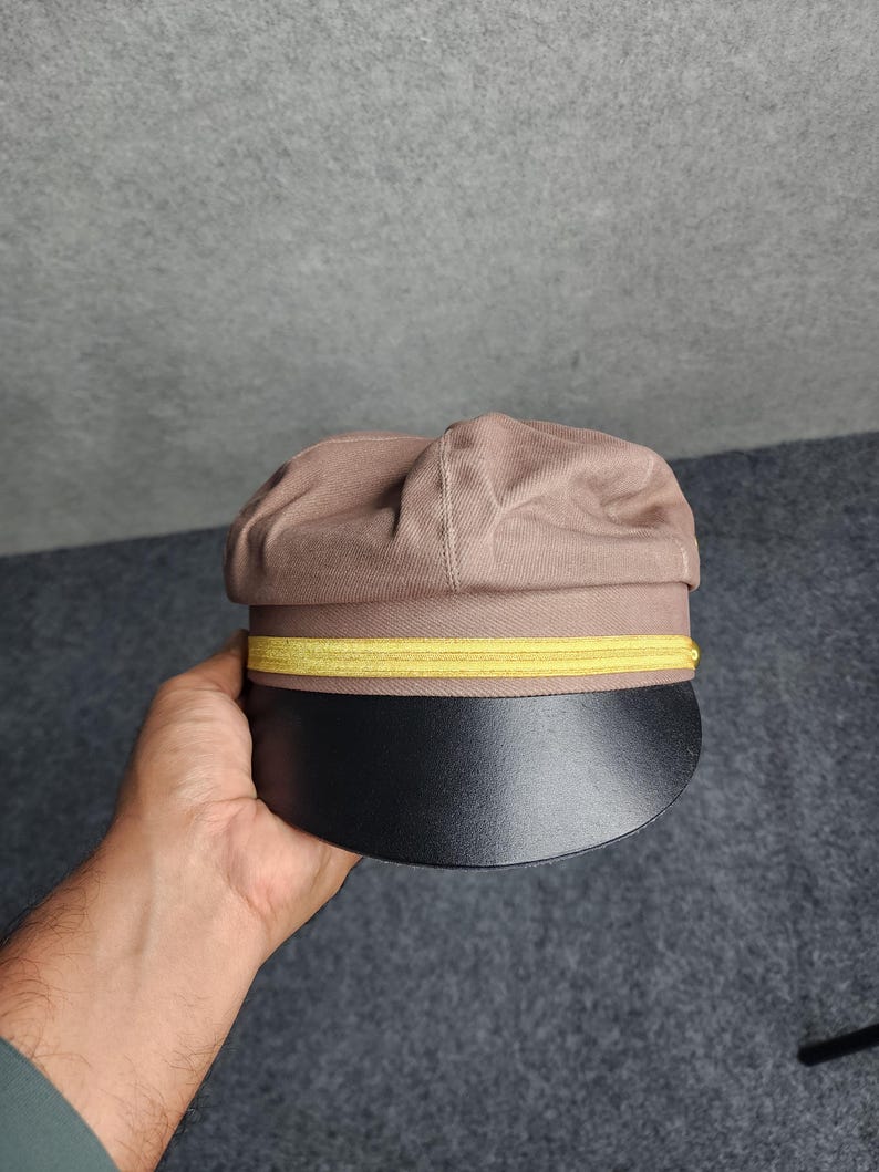 May include: A brown cap with a black brim and a yellow band. The cap is held in a hand against a gray background. The cap has a rounded top and a stiff, curved brim.