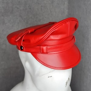 Puede incluir: Un sombrero de cuero rojo brillante con visera y correa. El sombrero se exhibe sobre una cabeza de maniquí blanca sobre un fondo gris. El sombrero tiene un acabado brillante.