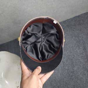 May include: A tan and black hat with a black interior. The hat has a brown trim and gold-colored buttons on the sides. The hat is held by a hand.