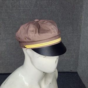 May include: A brown cap with a black brim and a gold-colored band. The cap has a rounded crown and a small button on top. The brim is a solid black color. The cap is displayed on a mannequin.