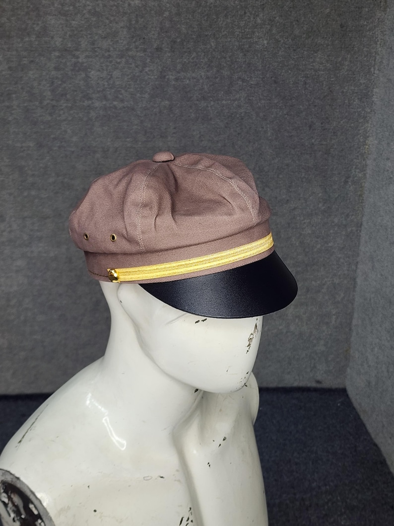 May include: A brown cap with a black brim and a gold-colored band. The cap has a rounded crown and a small button on top. The brim is a solid black color. The cap is displayed on a mannequin.