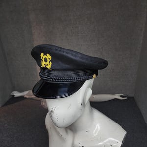 May include: Black captain's hat with a shiny black visor and a gold emblem. The hat features a black rope detail around the crown and small gold buttons. The hat is displayed on a white mannequin head.