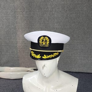 Könnte beinhalten: Eine weiß-schwarze Kapitänsmütze mit einem gold gestickten Ankeremblem auf der Vorderseite und dem Wort CAPTAIN darunter. Die Mütze verfügt über eine goldene Kordel am schwarzen Band und eine goldene Blattstickerei auf dem schwarzen Schirm.
