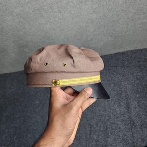 May include: A brown, military-style cap with a black brim and gold accents. The cap features a yellow band and two small eyelets on the crown. A gold button is on the side of the cap.