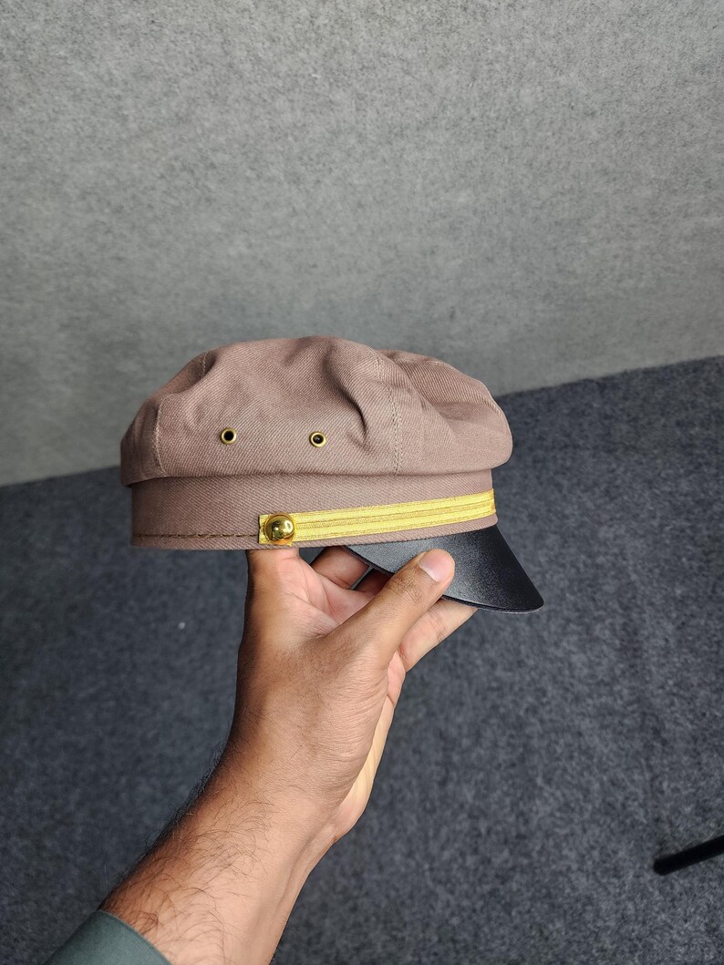 May include: A brown, military-style cap with a black brim and gold accents. The cap features a yellow band and two small eyelets on the crown. A gold button is on the side of the cap.