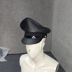 Op de afbeelding: Een zwarte leren hoed met een afgeronde kroon en een stijf, gebogen vizier. De hoed heeft een glanzende band en kleine goudkleurige accenten. De hoed wordt getoond op een witte mannequin.