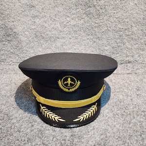 Könnte beinhalten: Schwarze Pilotenmütze mit glänzendem schwarzem Schirm und goldenem Emblem mit einem Flugzeug in einem Kranz. Die Mütze hat ein goldenes Band und goldene Blattakzente.