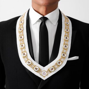 May include: A gold-toned chain collar with red and white emblems, worn over a white collar and black suit. The chain features repeating circular designs with a central emblem. The suit has a black tie and a white pocket square.