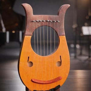 May include: A wooden lyre with a smiling face design. The instrument features a light brown body with a dark brown top section and a red-brown mouth. Six strings are visible, and the lyre stands on a small wooden base.