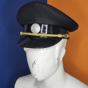 May include: Black hat with a shiny, patent leather-like brim and a circular silver emblem. The hat features a gold rope detail and is displayed on a white mannequin head. The background is split between blue and orange.