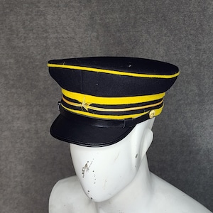 May include: A black and yellow military-style hat with a star emblem. The hat features a flat top with a yellow band and trim, a black visor, and a gold-colored button. The hat is displayed on a white mannequin head.