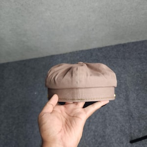 May include: A brown newsboy cap with a rounded crown and a stiff brim. The cap is held in a hand, showcasing its shape and texture. The cap has a classic design, suitable for various styles.