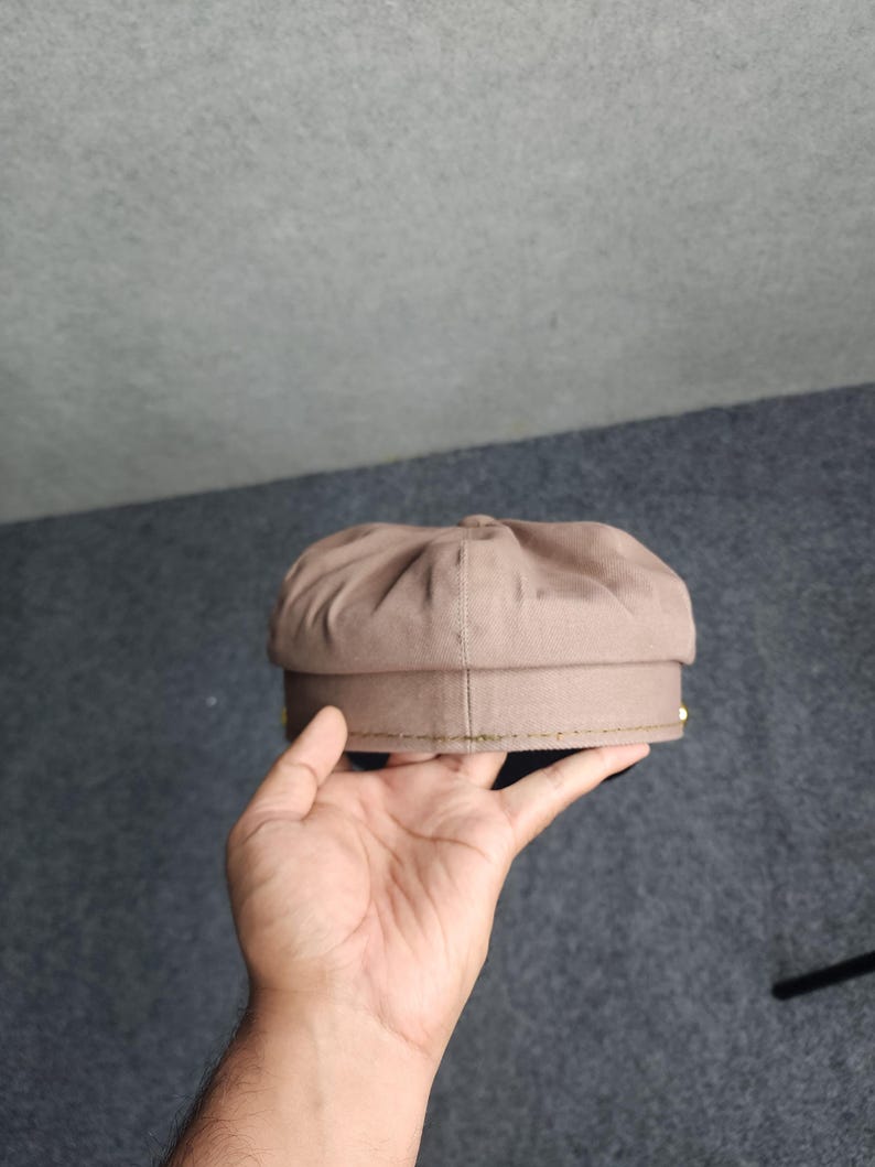 May include: A brown newsboy cap with a rounded crown and a stiff brim. The cap is held in a hand, showcasing its shape and texture. The cap has a classic design, suitable for various styles.