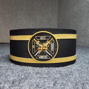 May include: A black ceremonial hat with gold trim and a circular emblem. The emblem features a crown, crossed swords, and the words "HOC SIGNO VINCES". The hat is likely used for formal events or as part of a uniform.