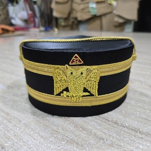 Puede incluir: Un sombrero negro y dorado con un emblema de águila bordado. El sombrero presenta adornos dorados y un pequeño emblema rojo y dorado. El sombrero es probablemente un tocado ceremonial o formal.