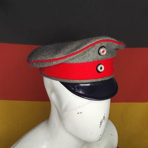 May include: A gray and red military hat with a black visor. The hat has a red band and trim, and two circular buttons on the front. The hat is displayed on a white mannequin head in front of a German flag.