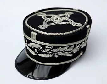 May include: A black hat with a glossy black peak. The hat is adorned with detailed silver embroidery, including a decorative knot and floral motifs. The hat's design is structured and rounded, with silver trim around the edges.