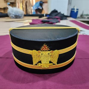 Puede incluir: Sombrero ceremonial negro con ribete dorado y un emblema bordado. El sombrero presenta un cordón dorado en el borde superior y una banda dorada en el centro. El emblema incluye un águila y el número 32.