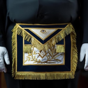 May include: A Masonic apron with a navy blue border and gold fringe. The apron features a white central panel with gold embroidery of wheat and the letter "G". Above the panel is a triangle with an eye. The person is wearing white gloves.
