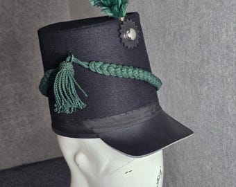 May include: A dark blue military hat with a black brim, adorned with a green feather, a green tassel, and a decorative silver-colored emblem. The hat is displayed on a white mannequin head.