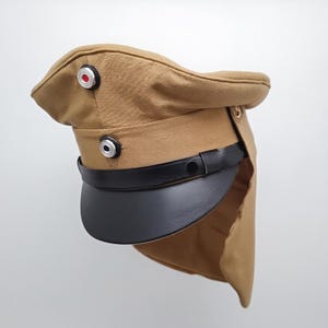 Puede incluir: Gorra de estilo militar de color beige con visera negra y correa de cuero negro. La gorra tiene dos botones plateados con centros rojos y negros. Tiene un ala curvada y una solapa que se extiende hacia atrás.