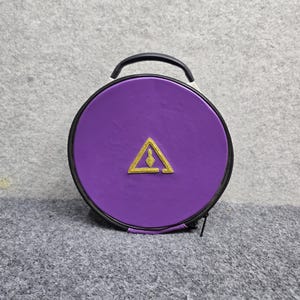 Op de afbeelding: Een ronde, paarse draagtas met ritssluiting en een zwart handvat. De voorkant van de tas is voorzien van een centraal, geborduurd goudkleurig driehoekig embleem met een kleine, puntige vorm erin. De tas heeft een zwarte rand en ritssluiting.