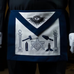 Puede incluir: Delantal masónico blanco y azul marino con símbolos bordados, incluyendo un ojo, columnas y herramientas. El delantal se lleva con un traje negro y guantes blancos. El diseño del delantal es simétrico y detallado.