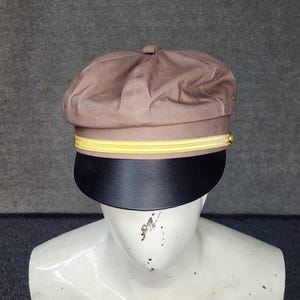 May include: A brown cap with a black brim and a yellow stripe. The cap is displayed on a white mannequin head. The cap has a rounded top and a stiff, curved brim.