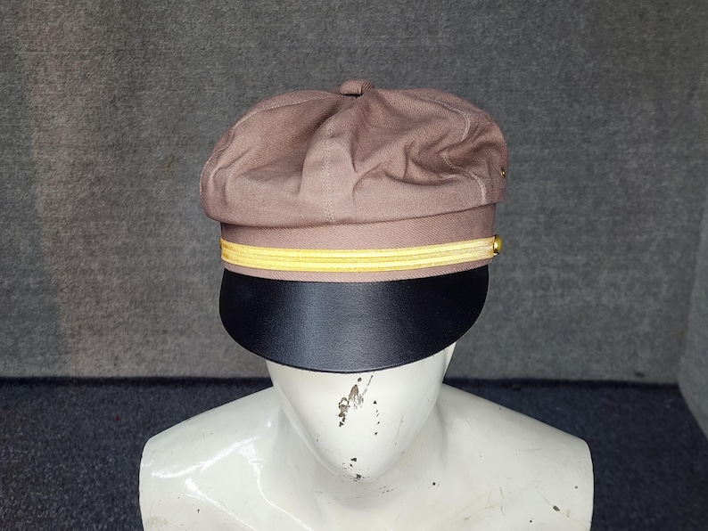 May include: A brown cap with a black brim and a yellow stripe. The cap is displayed on a white mannequin head. The cap has a rounded top and a stiff, curved brim.