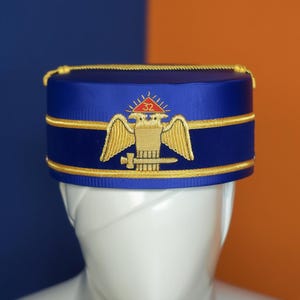 Puede incluir: Un sombrero azul con ribete dorado y un emblema bordado en oro con un águila, un cuadrado y el número 32. El sombrero se exhibe sobre una cabeza de maniquí blanca sobre un fondo azul y naranja.