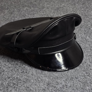 May include: A black leather hat with a shiny, patent leather brim. The hat features a structured crown and a strap detail. The hat is a classic style, suitable for various occasions.