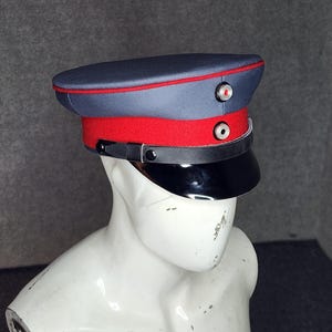 May include: A vintage-style hat with a dark blue crown, a red band, and a black visor. The hat features a red trim and two decorative buttons. The hat is displayed on a white mannequin head.