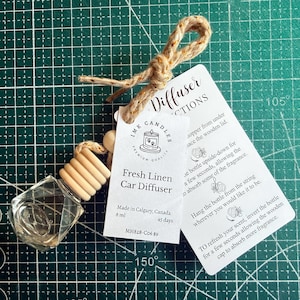 May include: A clear glass car diffuser bottle with a faceted shape and a light wood cap, suspended by a tan jute twine. Attached is a white rectangular tag labelled Fresh Linen Car Diffuser by IMK Candles, including 8 ml volume text.