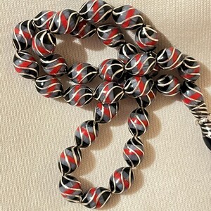 Peut inclure: Un collier de perles avec des perles ovales à motif spiralé rouge, noir et gris. Les perles sont séparées par des accents métalliques argentés. Le collier est présenté sur une surface de couleur neutre.