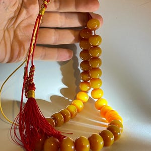 May include: A necklace with large, round, amber-coloured beads. The beads change colour from a deep amber to a bright orange. The necklace has a red tassel and a braided red and gold cord.