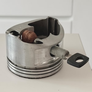 Puede incluir: Un pistón de metal reutilizado con una nuez en el interior, asegurado por un perno roscado y un mango negro en forma de llave. El pistón tiene un acabado plateado y está sobre una superficie blanca.