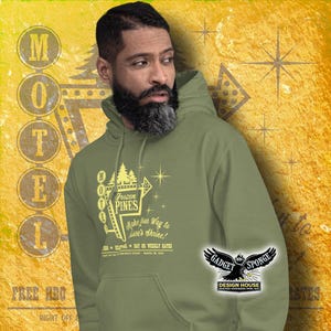 Może przedstawiać: Zielona bluza z kapturem z grafiką w stylu vintage przedstawiającą znak motelu z napisem "Frozen Pines" oraz tekstem "Make your way to Stan's Shrine!" i "Day or Weekly Rates". Grafika jest w kolorze ż&oacute;łtym i przedstawia stylizowaną sosnę. Bluza ma czarno-białe logo "Gadget Sponge Design House" w prawym dolnym rogu.