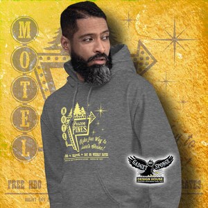 Może przedstawiać: Szara bluza z kapturem z ż&oacute;łtym nadrukiem przedstawiającym vintage'owy znak motelu z napisem "Frozen Pines Motel" oraz tekstem "Make your way to Love's Shrine!" i "Day or Weekly Rates". Nadruk zawiera r&oacute;wnież gwiazdę i sosnę. Bluza ma czarno-białe logo na rękawie z napisem "Gadget Sponge Design House".