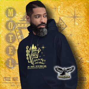 Może przedstawiać: Męska bluza z kapturem w kolorze granatowym z ż&oacute;łtym nadrukiem przedstawiającym vintage'owy znak motelu z napisem "Frozen Pines Motel" i "Make your way to Lare's Shrine!" Znak ma ż&oacute;łtą gwiazdę i ż&oacute;łtą strzałkę wskazującą w prawo. Bluza ma białe logo w prawym dolnym rogu z napisem "Gadget Sponge Design House".