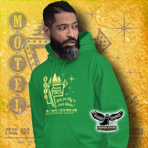 Może przedstawiać: Zielona bluza z kapturem z grafiką vintage'owego znaku motelu z tekstem "Frozen Pines" i "Make your way to Lave's Shrine!" Znak jest ż&oacute;łty z białą obw&oacute;dką i gwiazdami. Tekst "Free HBO Right Off" jest widoczny w lewym dolnym rogu obrazu.