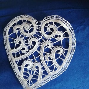 May include: A white, heart-shaped lace doily with intricate, swirling patterns. The delicate lace is set against a blue background, showcasing the detailed craftsmanship. The doily's edges are finely scalloped, adding to its elegant appearance.
