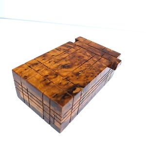 Peut inclure: Une boîte à puzzle en bois de forme rectangulaire. La boîte est faite de différents grains de bois, avec un mélange de tons bruns clairs et foncés. Le dessus de la boîte présente une série de pièces imbriquées.