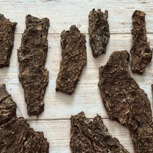 May include: A collection of irregularly shaped pieces of tree bark, varying in size and texture. The bark is primarily brown with lighter and darker shades, and some pieces have a rough, textured surface. The pieces are arranged on a light-colored wooden surface.