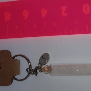 May include: A silver key chain with a small glass bottle filled with white glitter. The bottle has a cork stopper and a silver charm that says "Wish". The keychain is attached to a brown card that says "Handmade with love".