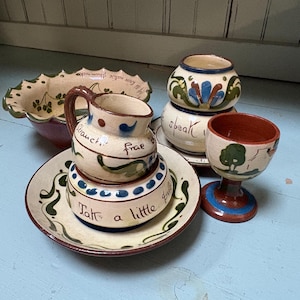 May include: A collection of hand-painted ceramic pottery with a rustic aesthetic. The set includes a bowl, a small pitcher, a plate, a small jar, and an egg cup. The pieces feature floral and script designs in shades of brown, green, blue, and red.