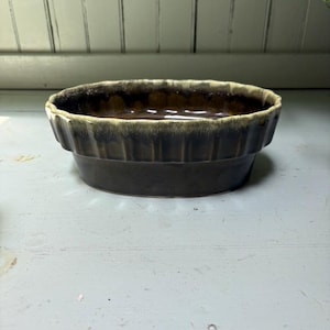 May include: An oval ceramic bowl featuring a deep brown glaze with a lighter cream-colored drip effect along the scalloped rim. The exterior of the bowl has a vertical ribbed texture, while the interior is smooth and dark brown in color.